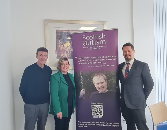Image shows three people smiling, indoors, Scottish Autism supporter banner