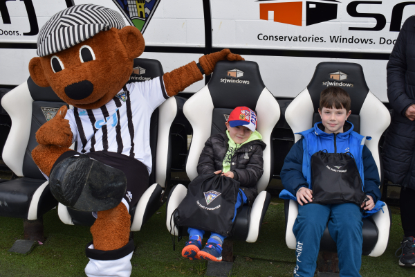 Photograph shows two young boys sitting next to a football mascot in a football dugout