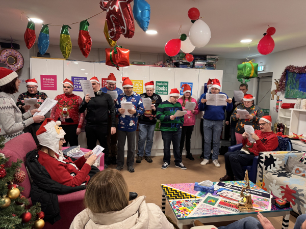 An image of a choir, wearing festive jumpers and hats, singing indoors in an art studio