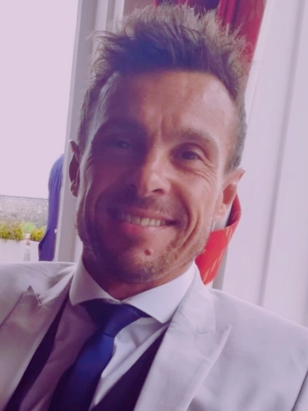 A man in a white suit and tie smiles towards the camera