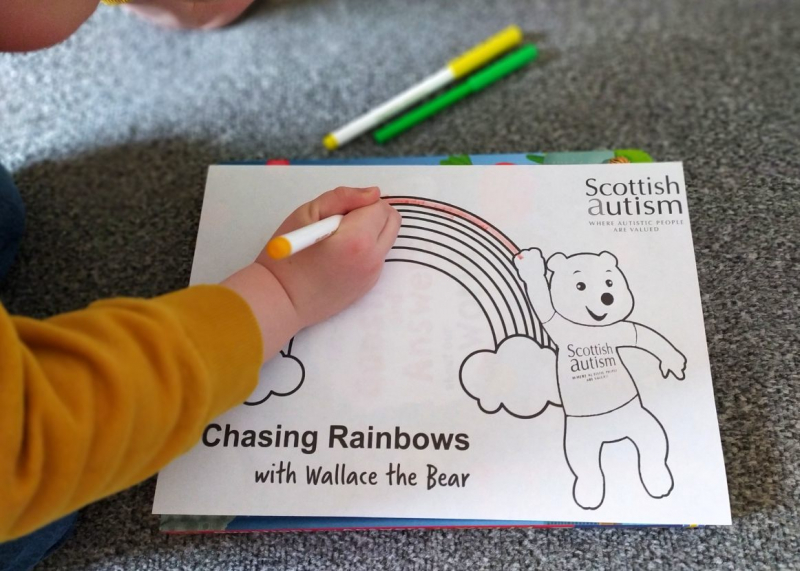 Child colours in the rainbow of a colouring sheet with a rainbow and Scottish Autism mascot bear on it
