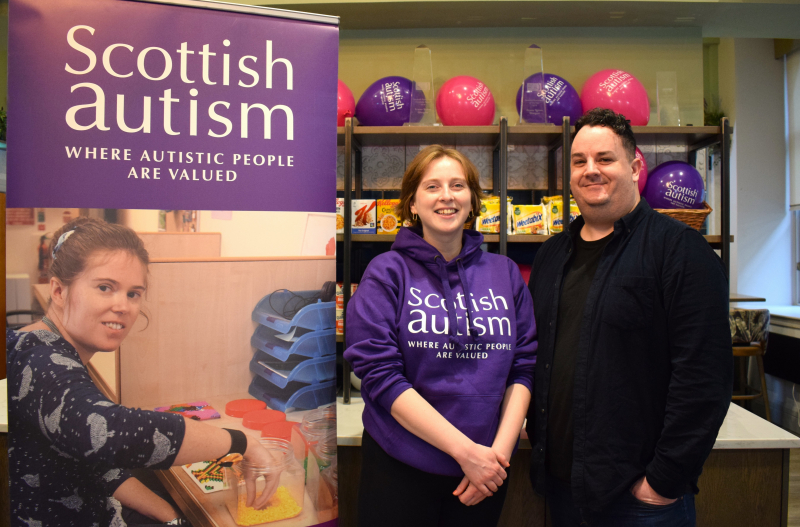 Image is of two people launching a charity partnership, hotel breakfast room, Scottish Autism pop up banner, Text reads "Scottish Autism, Where autistic people are valued"