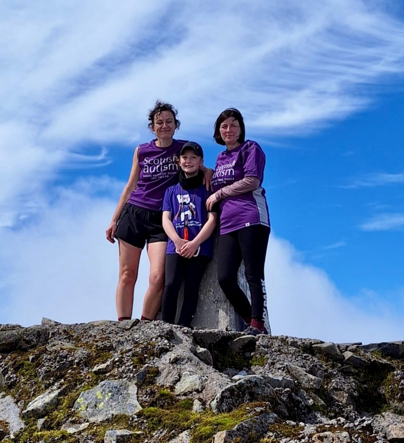 Image of two adults and a child, wearing Scottish Autism supporter tshirts, standing at the summit of a mountain with a blue sky background