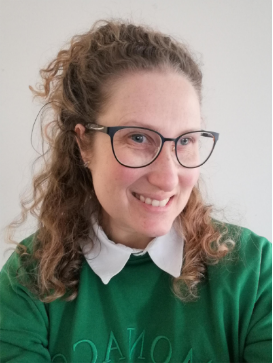 A person with long wavy hair smiles for the camera, wearing a green jumper