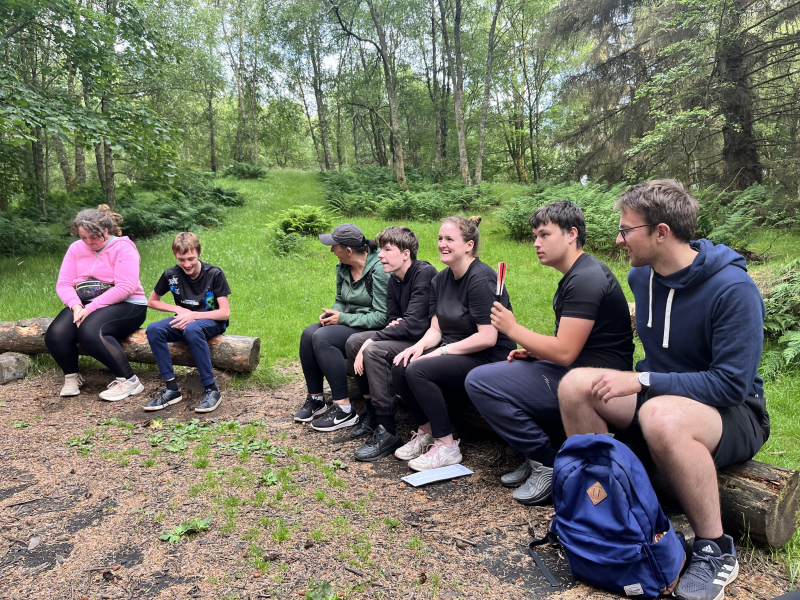 seven people sitting on a log outdoors, smiling and talking