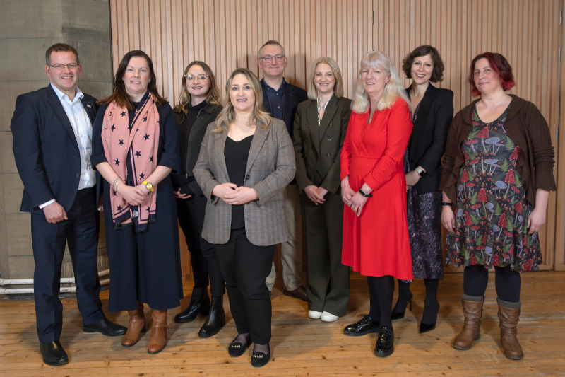 A group photo of nine individuals standing in front of a wooden paneled wall. They are all dressed in business or smart casual attire at Scottish Autism's Centre launch, smiling at the camera.