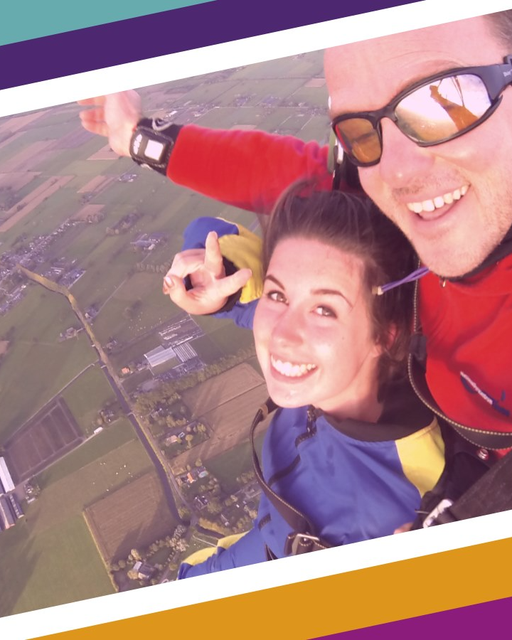 Two people smile at the camera as the skydive in tandem with an ariel view of the ground