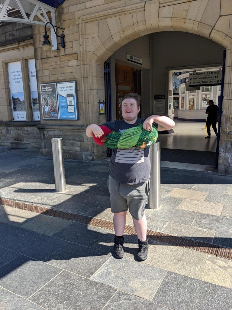 Young adult standing outside train station, smiling holding the hungry caterpillar soft toy