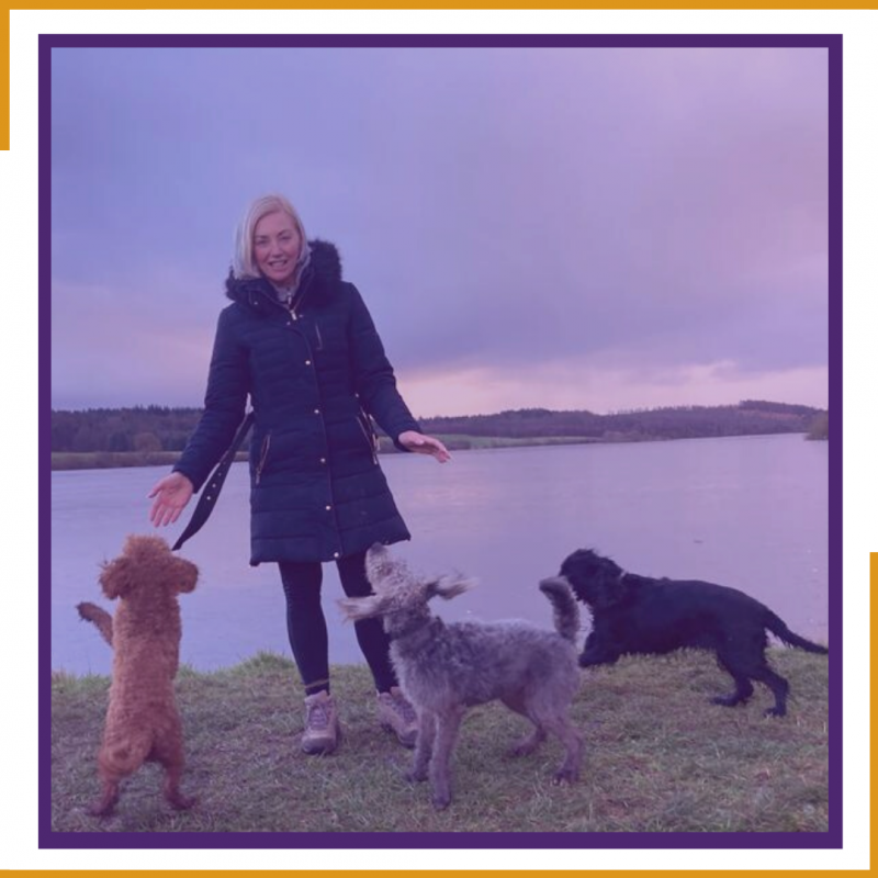 A woman stands infront of a loch while three dogs run around at her feet