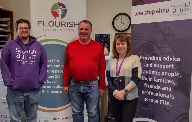 Image is of three people standing in a room, smiling for the camera, with pop up banners promoting Scottish Autism's Fife One Stop Shop and Flourish
