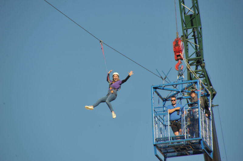 Image is of a person on a zipline, next to a crane, sky background