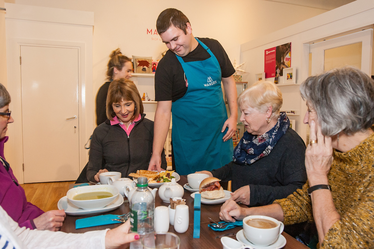 Four ladies sat around a table with food and drink in front of them being served by a young man in a black t-shirt and teal apron
