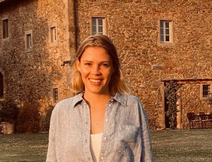 A woman with a light shirt on and blonde hair smiles as she stands outside an traditional looking building