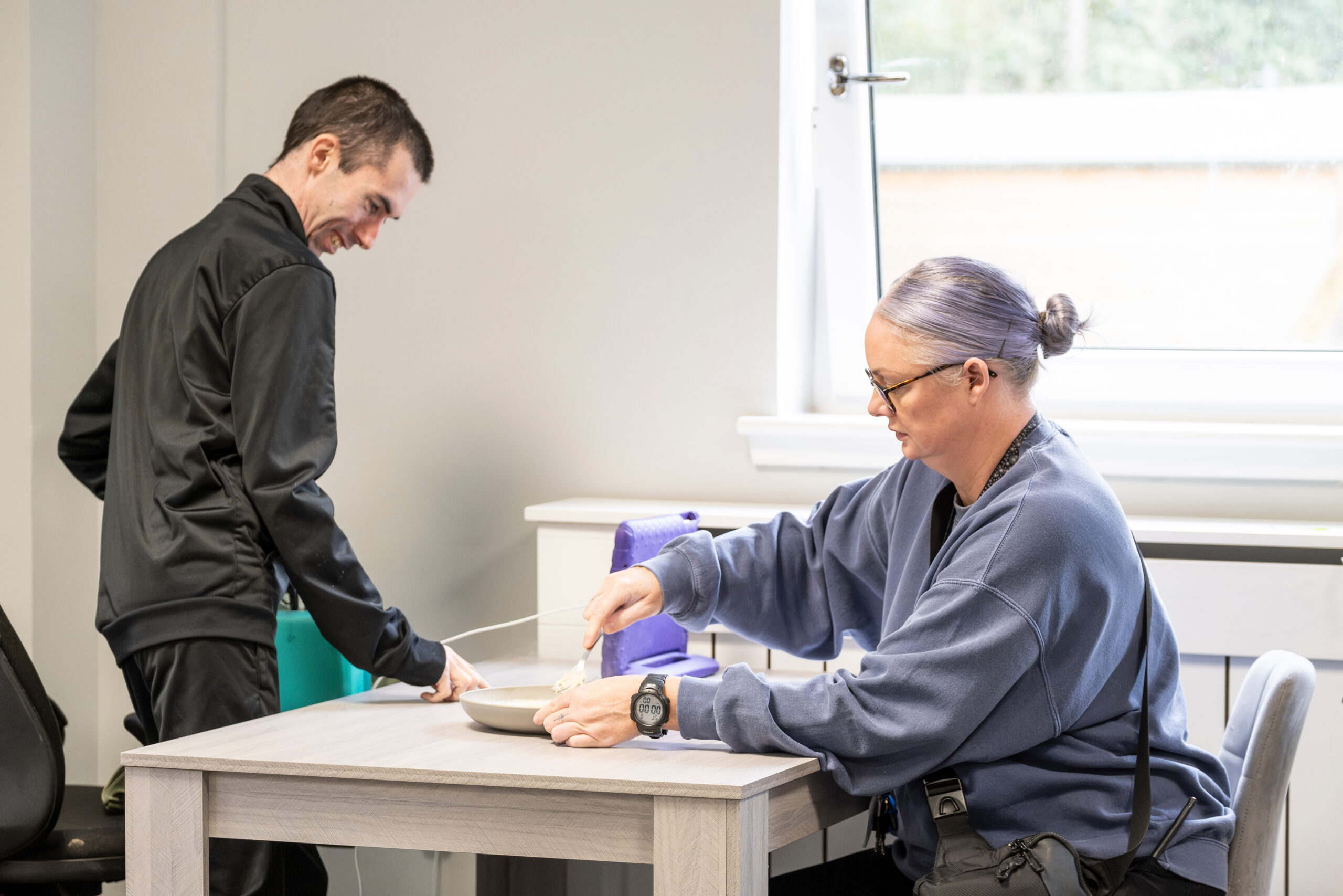 Image is of two people sitting down to eat together at a table. One person is smiling while the other is putting something in a bowl