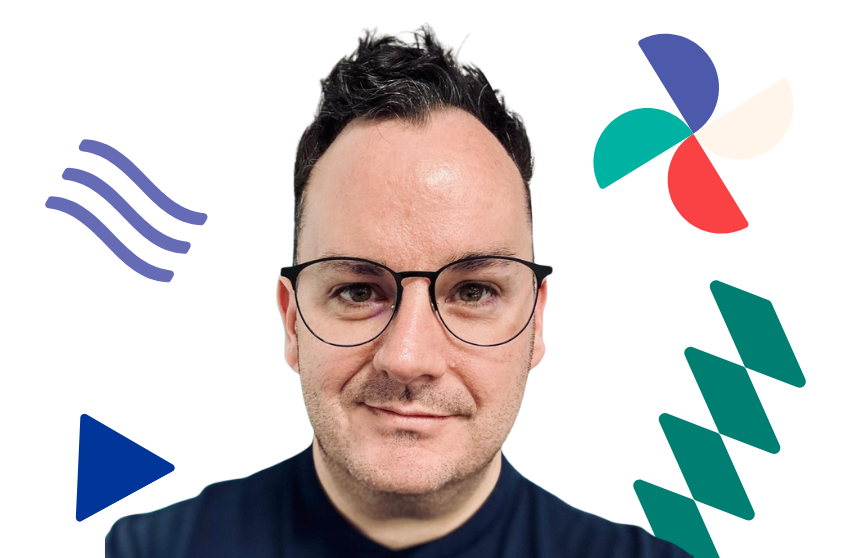 A man with glasses and a dark top and dark hair looks at the camera, colourful shapes are in the background