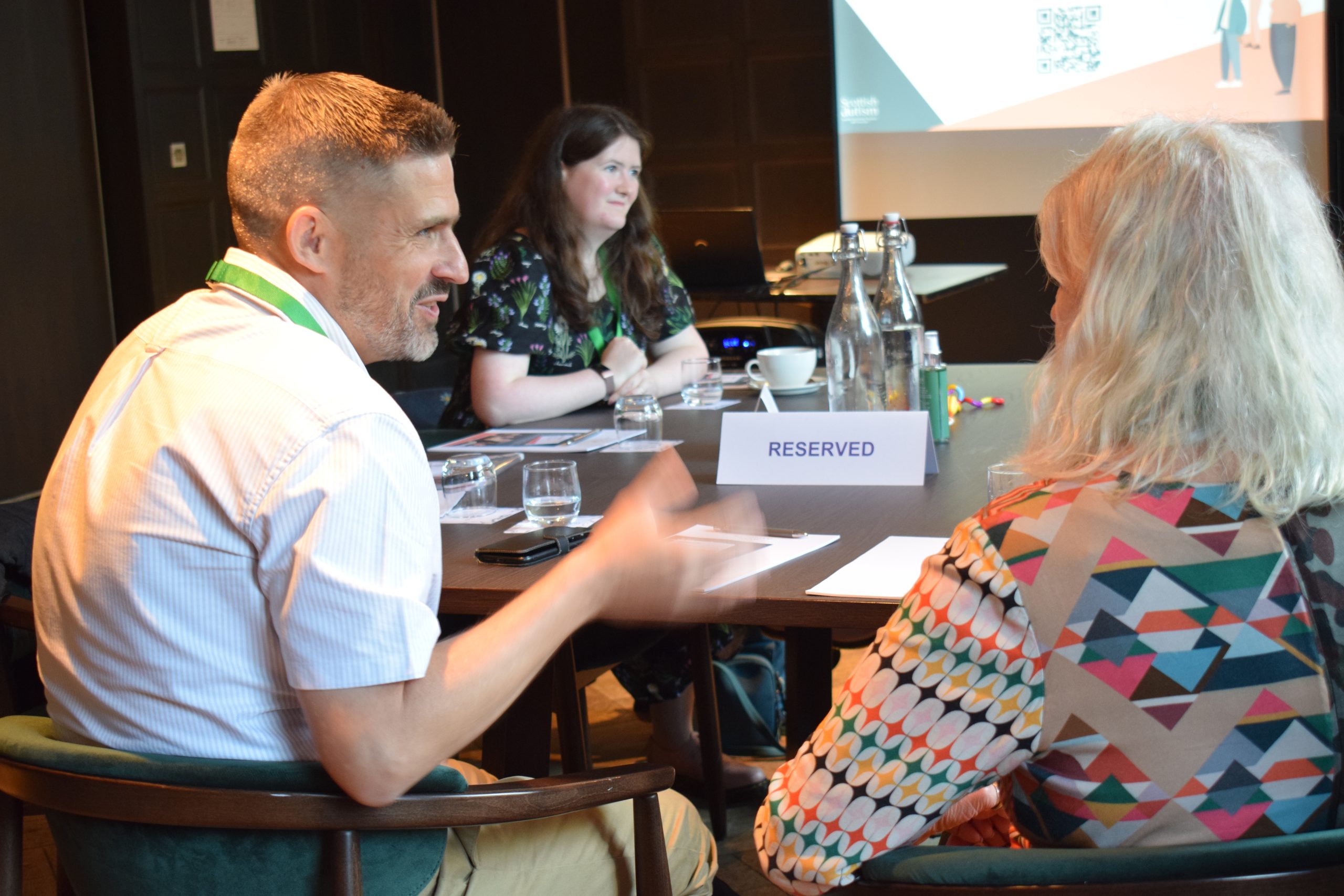 Three business people sit talking at an event with a presentation in the background