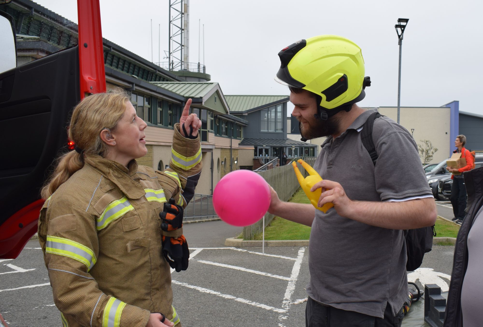 A female firefighter stands on the left of the photo and speaks to a young man in a grey t-shirt wearing a yellow firefighter's helmet