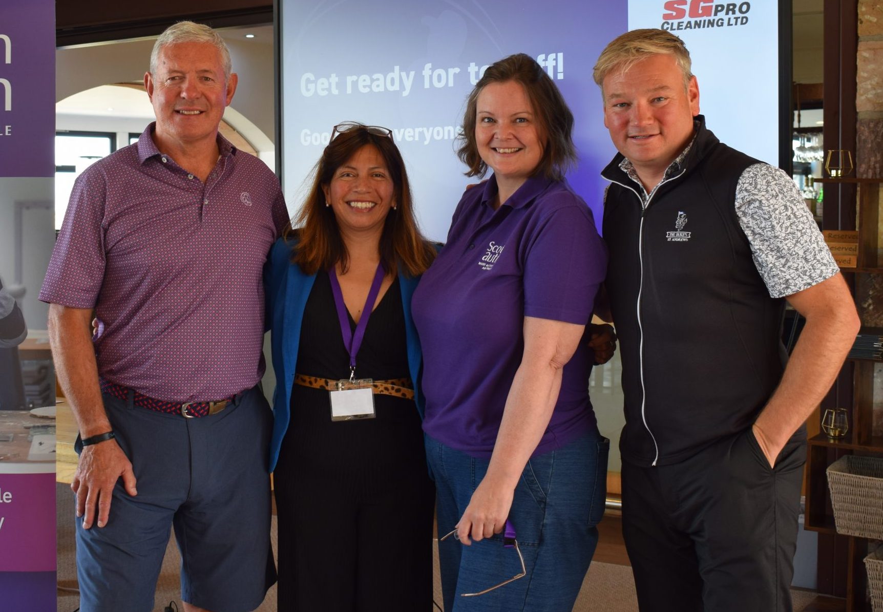 A group of four people stand together smiling for the camera at a golf event, with a presentation displayed in the background