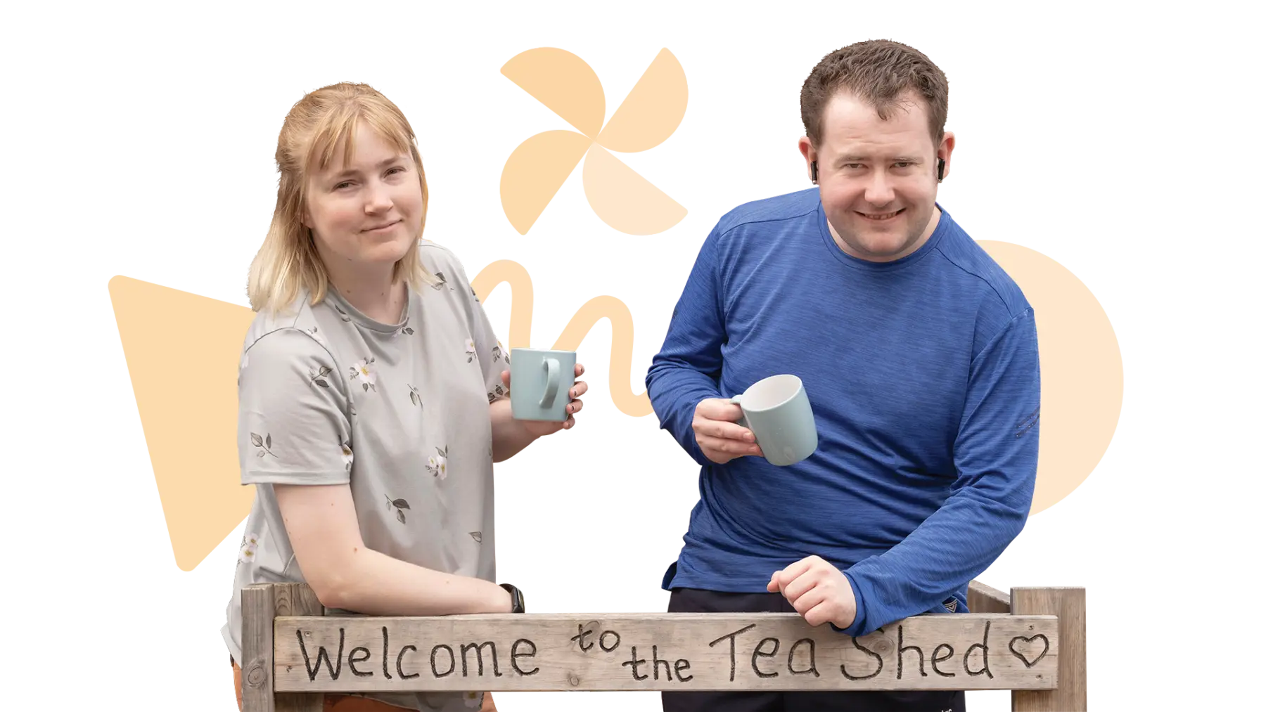 Image is of two people standing holding cups of tea, smiling at the camera, a sign is in front of them which says "Welcome to the Tea Shed"