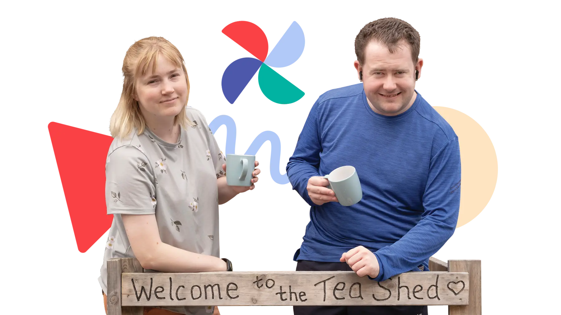 Image is of two people standing holding cups of tea, smiling at the camera, a sign is in front of them which says "Welcome to the Tea Shed"