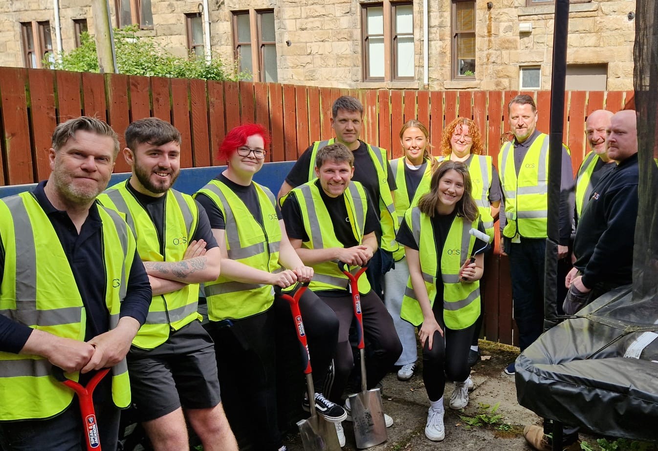 Image is of a group of 11 people wearing high vis vests, smiling for the camera, holding spades and garden work equipment