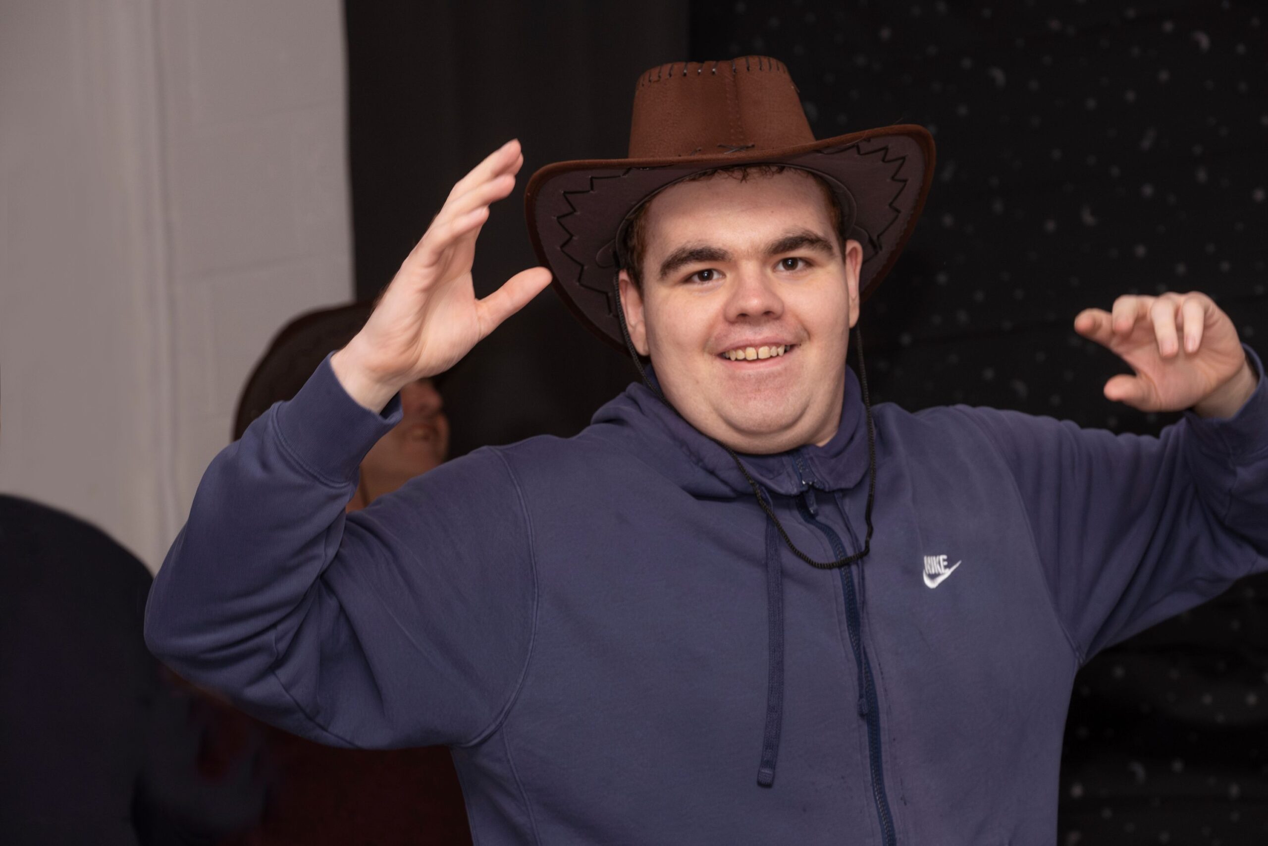 Young man in dark blue sweatshirt wearing brown cowboy hat smiling at the camera with his hands up in the air.