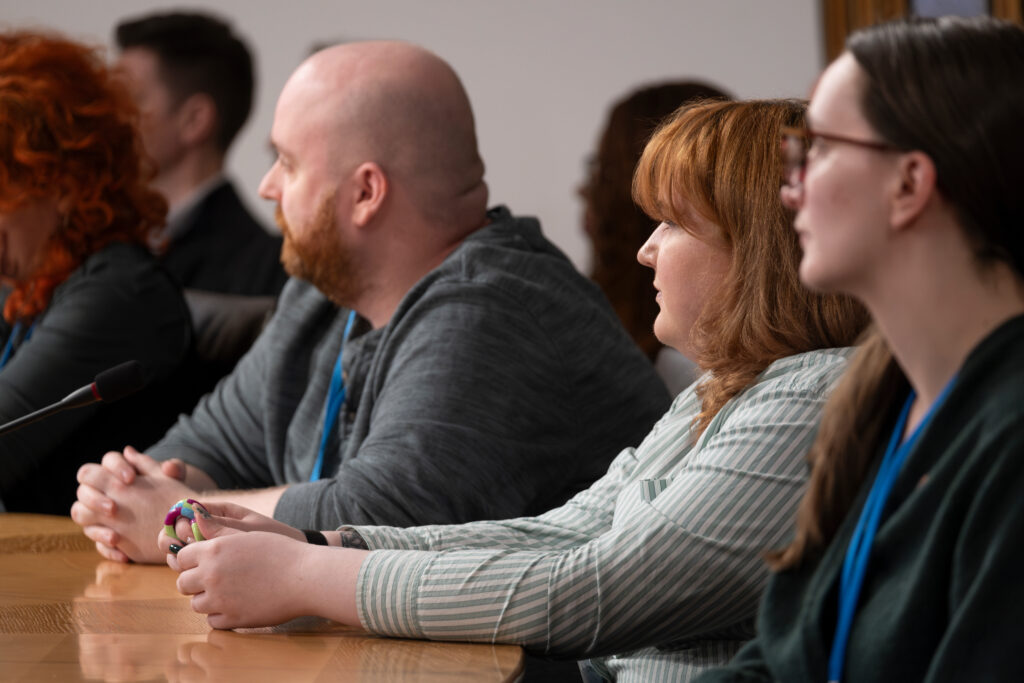 Image is a group of three people sitting listening to someone speaking at parliament