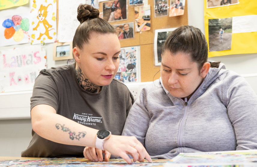 Two adult women sitting at a table whilst concentrating on a puzzle together