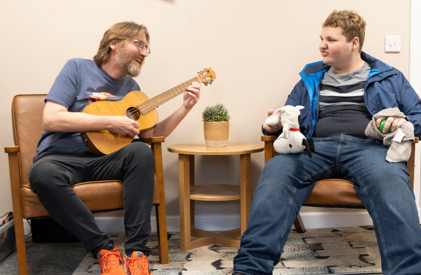 Two adults sitting on chairs facing each other smiling and talking while one plays guitar