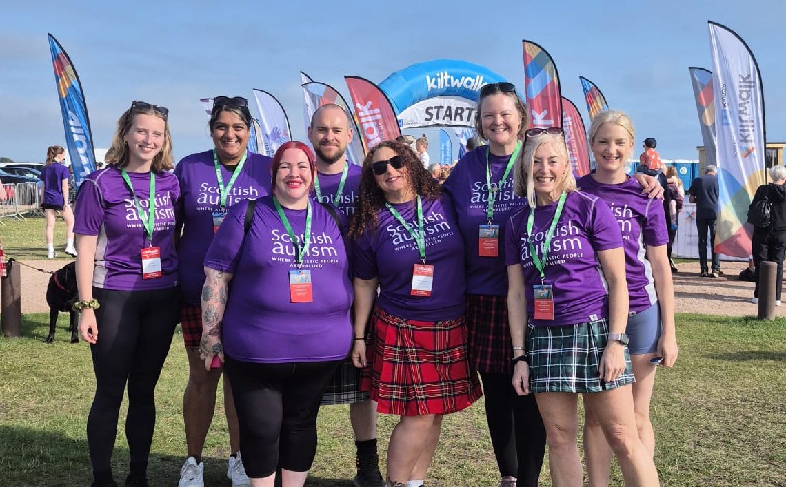 A group of eight Scottish Autism supporters pose for a photo at a kiltwalk event, wearing kilts and shorts