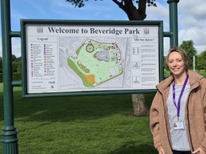 A woman smiling standing beside a park sign that reads "Welcome to Beveridge Park"
