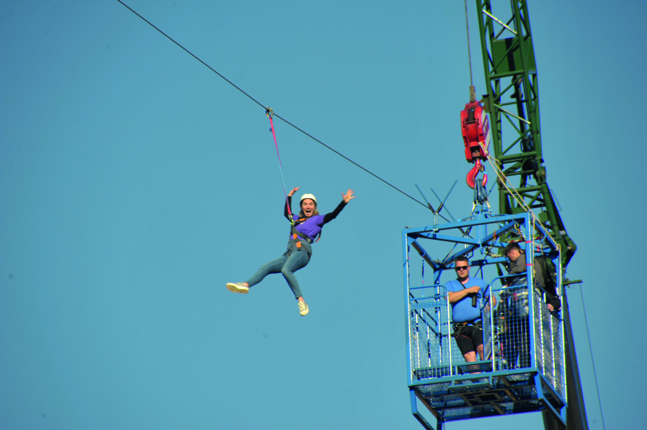 An image of someone zipsliding high up in the sky from a crane, with their hands in the air