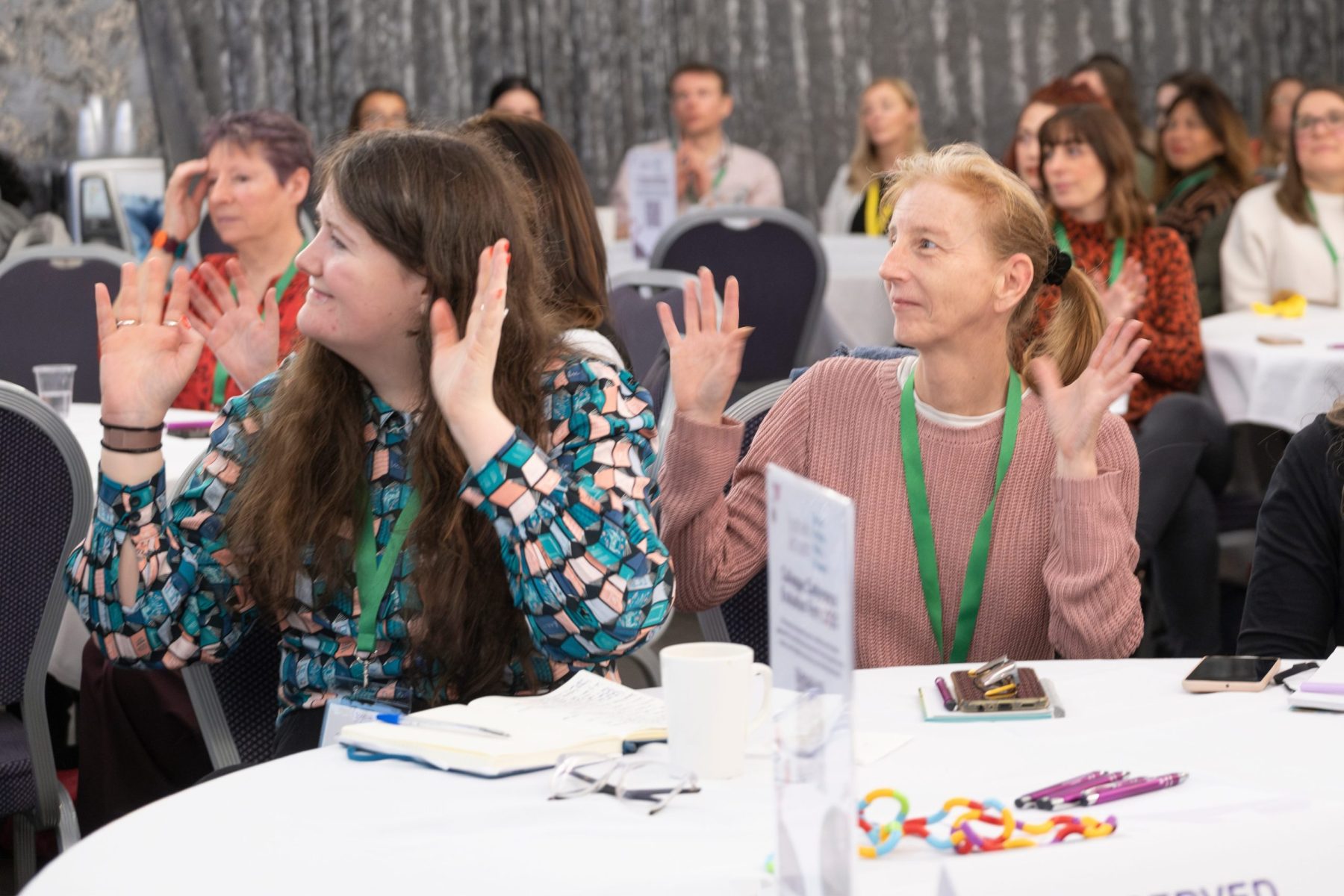 Room of people at a speaking event, two people in foreground raising hands in silent applause