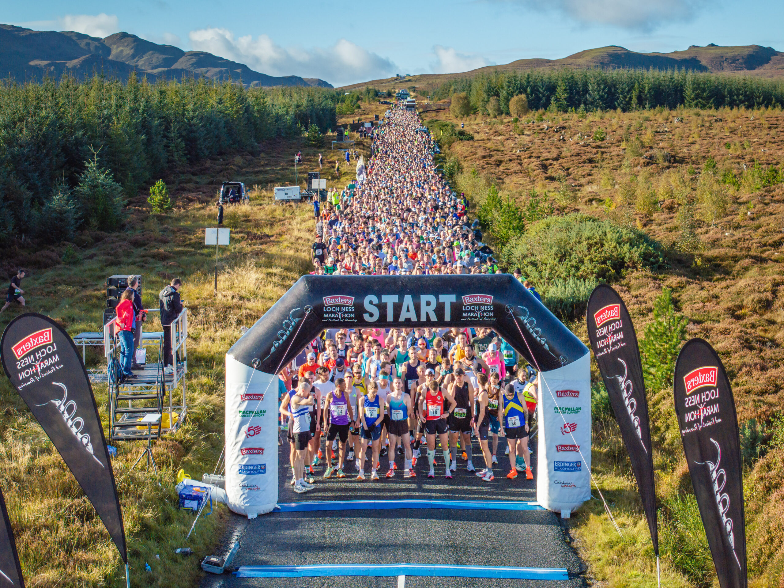 Image is of a starting line of runners before the marathon begins, in a green and hilly landscape