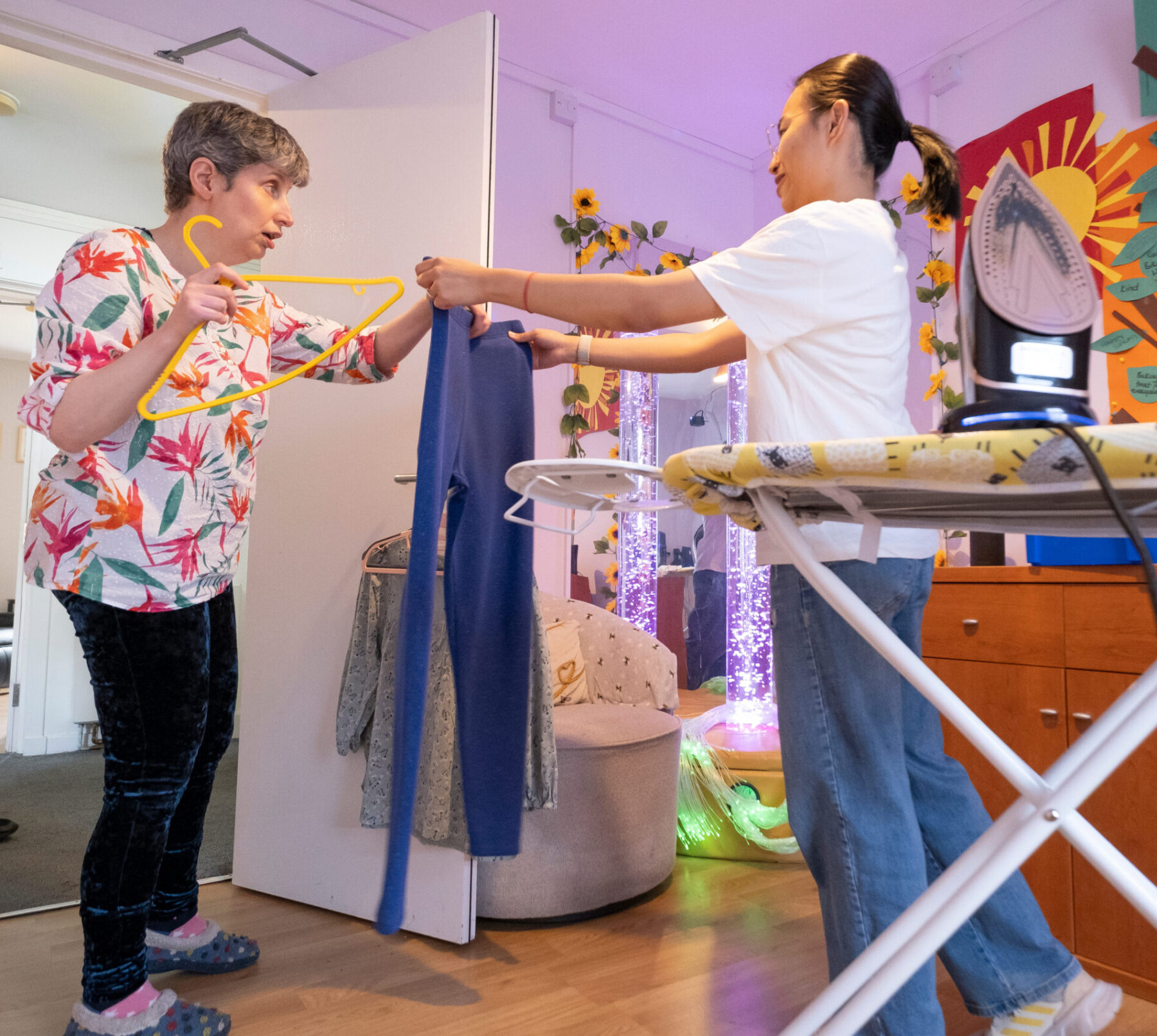 Two Ladies in a room. One is stood in front of an ironing board handing an item of clothing to another lady.