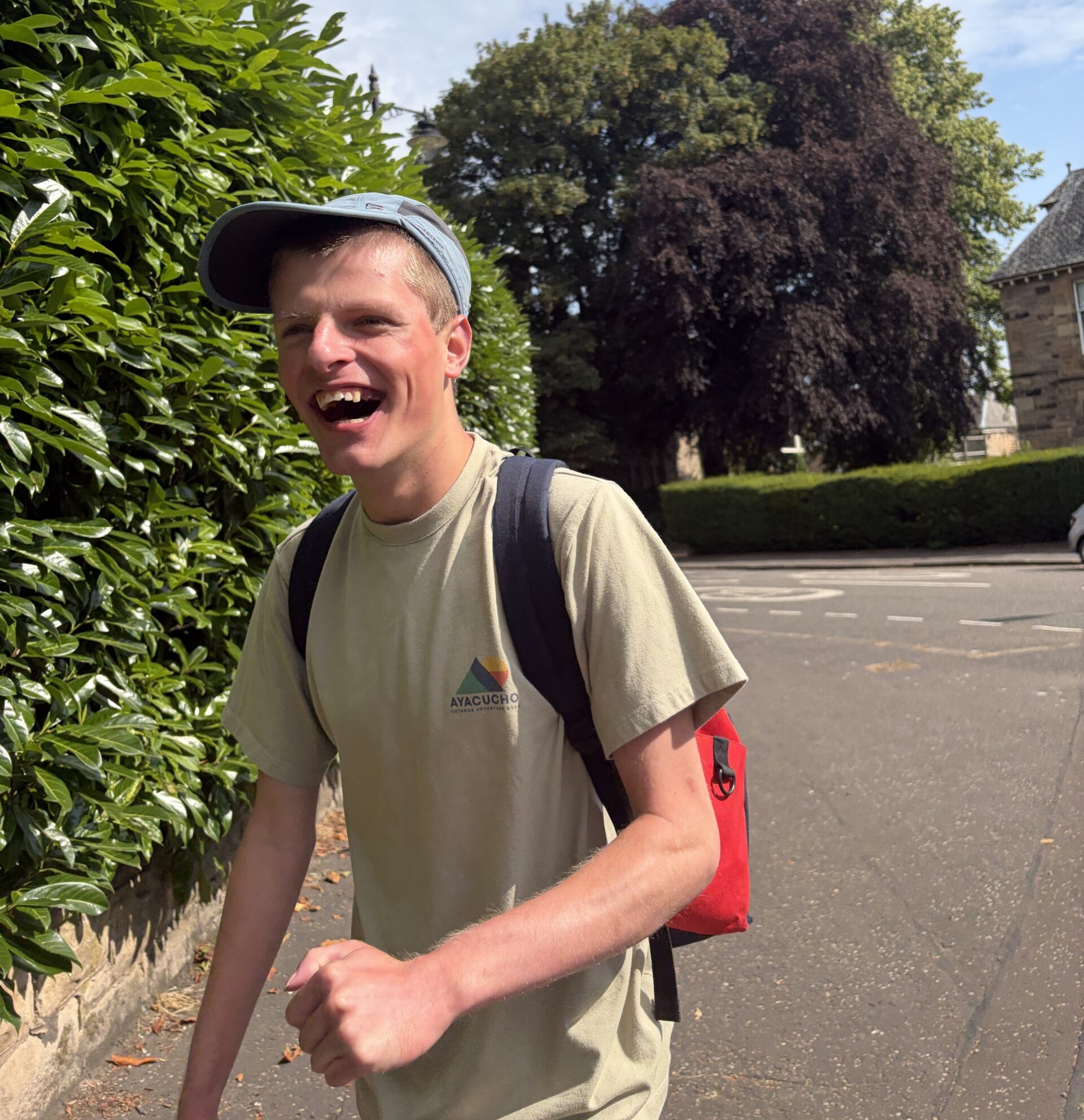 Image of a young man in a tan t-shirt and baseball cap walkign down the street laughing.