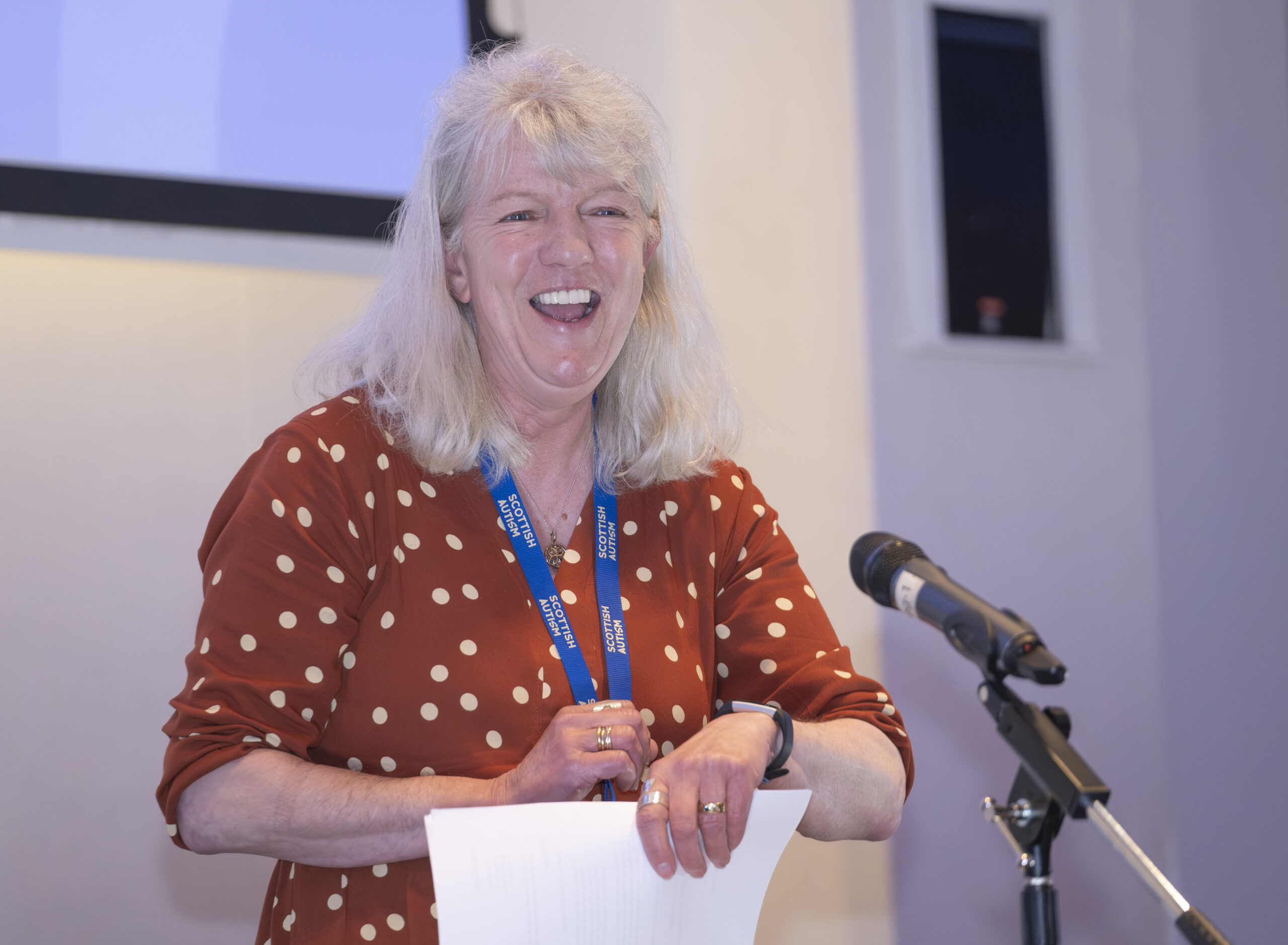 Scottish Autism Cheif Executive Dory McLaughlin stood in front of a mircophone at the Scottish Autism Colleague Conference, smiling and wearing a red dress with white spots.